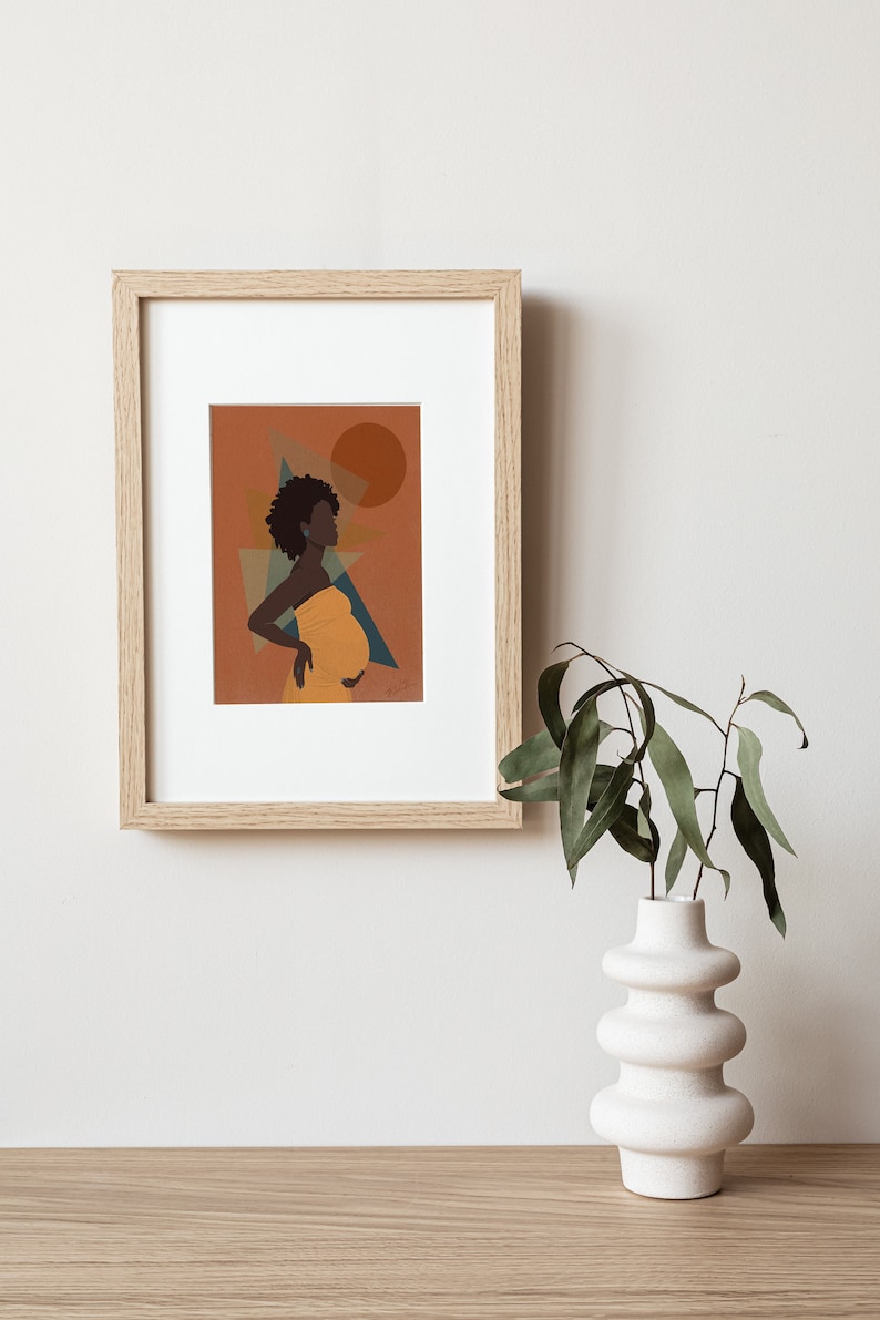 Op de afbeelding: Ingelijste kunstdruk met een silhouet van een zwangere persoon in een gele jurk, tegen een oranje achtergrond met geometrische vormen en een zon. Het kunstwerk bevindt zich in een licht houten lijst, tentoongesteld aan een witte muur. Een witte vaas met groene bladeren staat op een houten tafel.
