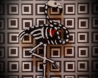 May include: A whimsical flamingo figurine with a skeleton design. The black and white body features bone details, a red heart, and a white head. The flamingo stands on one leg against a patterned background.