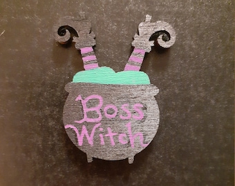 May include: Black wooden cauldron with green and purple paint. The cauldron has two witch legs sticking out of the top and the words "Boss Witch" written in purple paint.