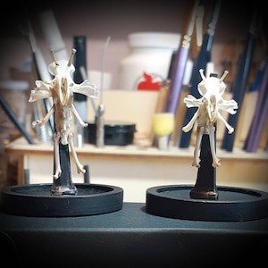 May include: Two small white skeletal figures on black bases. The figures are posed with their arms outstretched and their heads tilted back. The figures are on a dark surface.