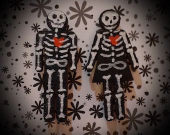 Puede incluir: Dos figuras de esqueleto de madera con corazones rojos pintados en el pecho. Las figuras están pintadas de negro y blanco y tienen un diseño simple.