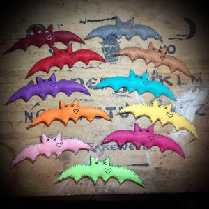 May include: A collection of ten felt bats in various colors, including red, orange, yellow, green, blue, purple, pink, and brown. Each bat has a stitched-on face with a heart and x-shaped eyes.