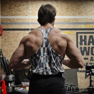May include: A person wearing a black tank top with a geometric pattern. The tank top has a racerback style. The person is standing in a gym with weights and other equipment in the background.