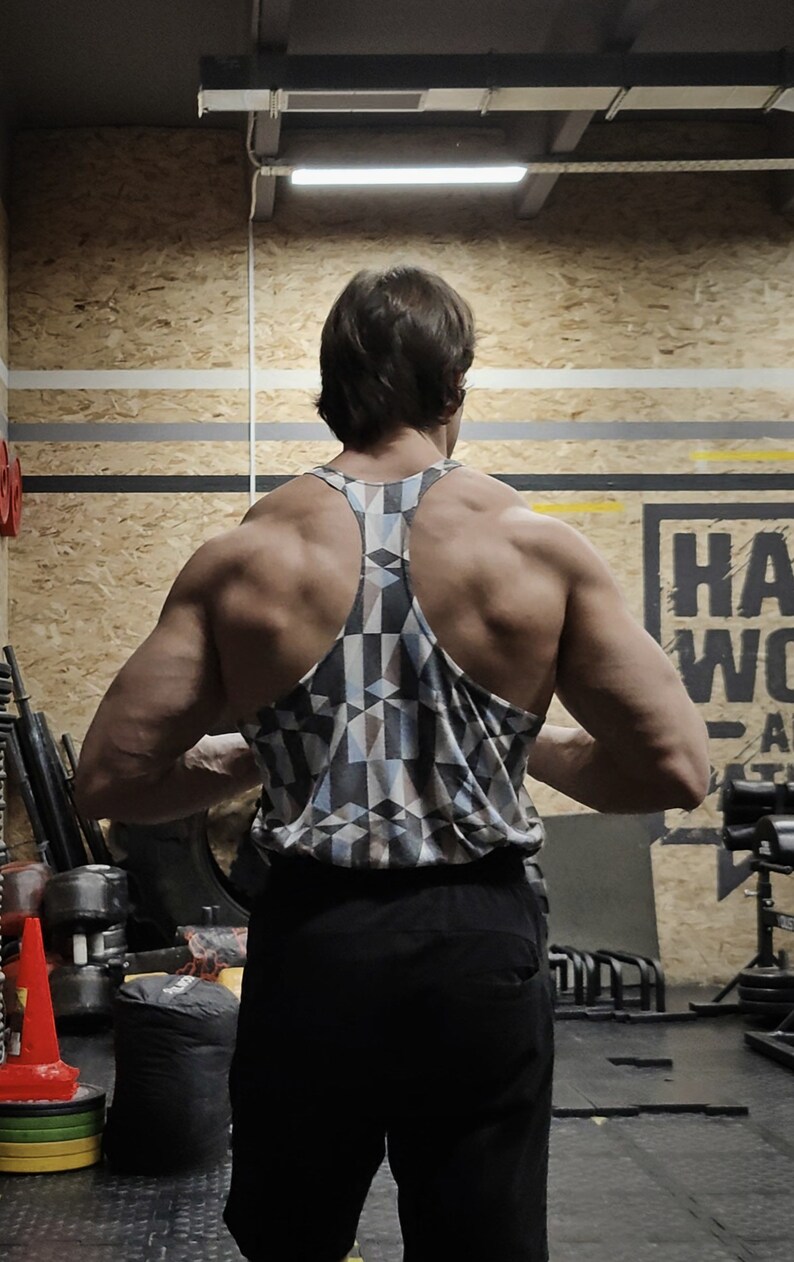 May include: A person wearing a black tank top with a geometric pattern. The tank top has a racerback style. The person is standing in a gym with weights and other equipment in the background.