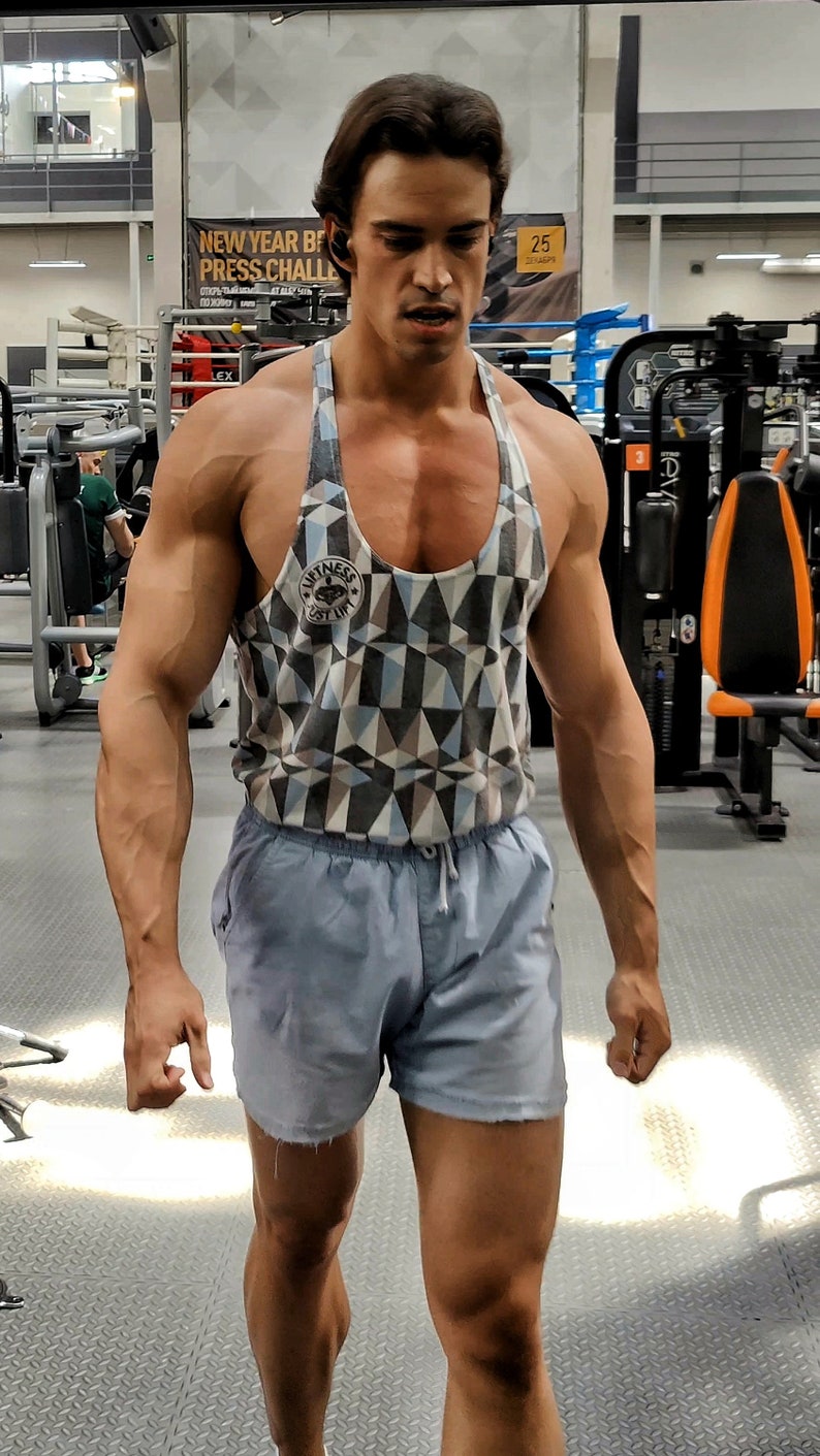 May include: A man wearing a gray and blue geometric patterned tank top with a black logo that says "Fitness First" and blue athletic shorts. He is standing in a gym.