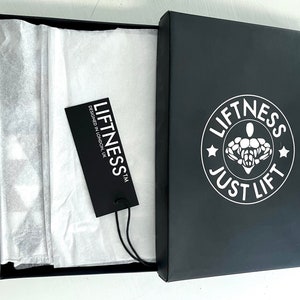 May include: A black box with a white logo that says "Liftness Just Lift" and a white star on each side of the logo. The box is open and there is a white tissue paper inside. A black tag with white text that says "Liftness TM Designed in London UK" is on top of the tissue paper.