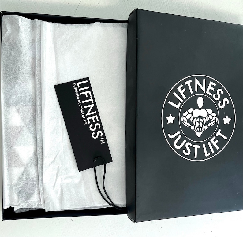 May include: A black box with a white logo that says "Liftness Just Lift" and a white star on each side of the logo. The box is open and there is a white tissue paper inside. A black tag with white text that says "Liftness TM Designed in London UK" is on top of the tissue paper.