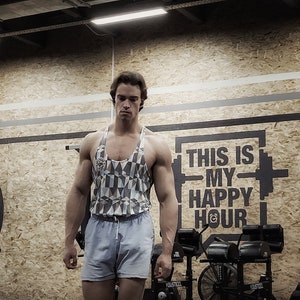 May include: A man wearing a white and gray patterned tank top and light blue shorts stands in a gym. The gym has a sign that says "This is my happy hour".