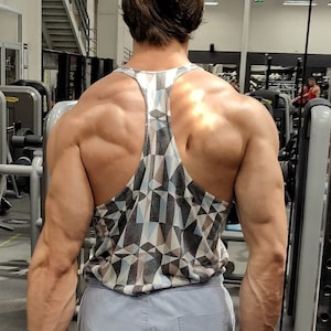 May include: A man wearing a gray and white geometric patterned tank top and gray shorts. He is standing in a gym with weightlifting equipment in the background.