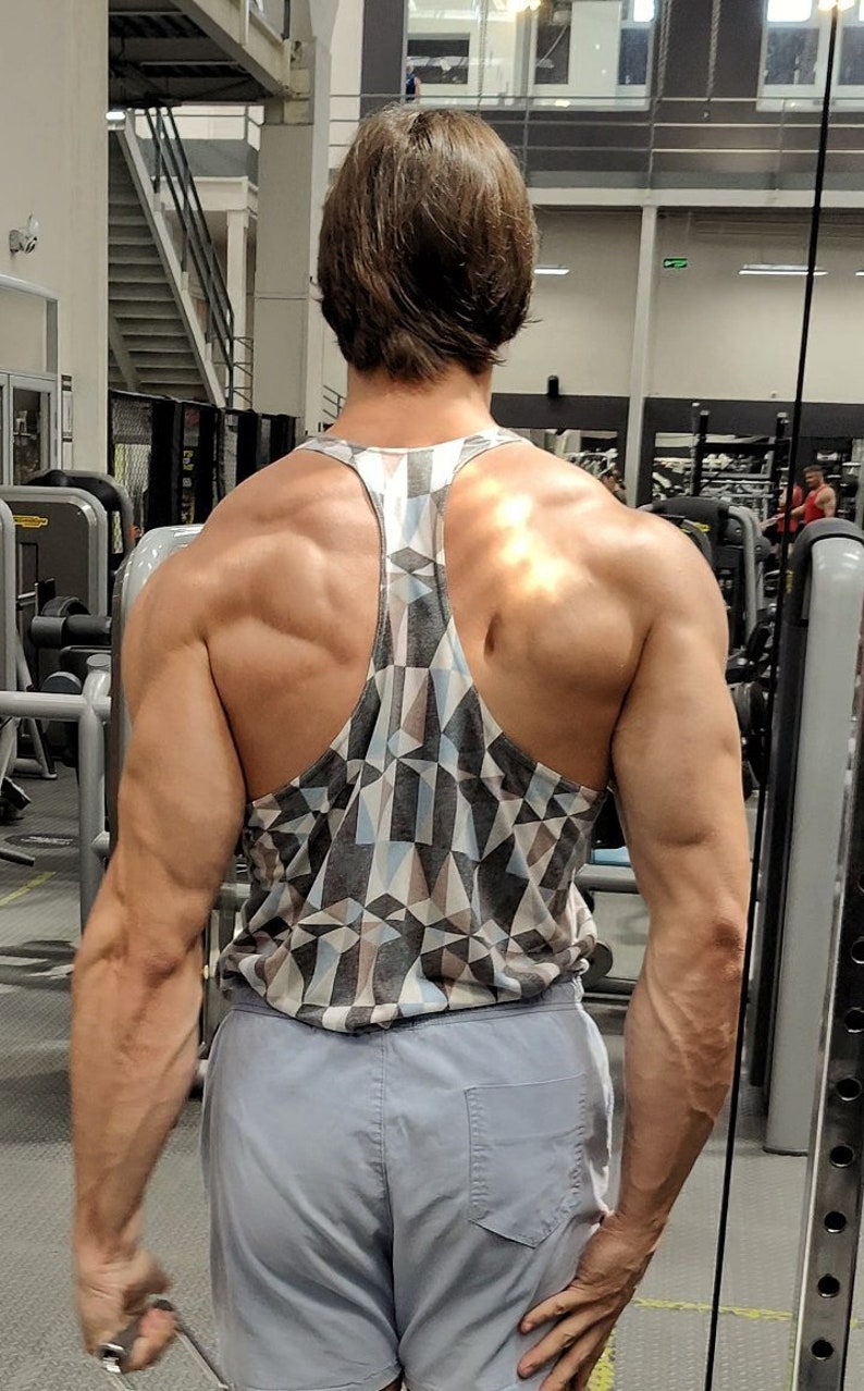 May include: A man wearing a gray and white geometric patterned tank top and gray shorts. He is standing in a gym with weightlifting equipment in the background.