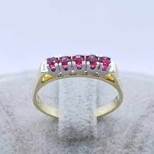 Könnte beinhalten: Ein Goldring mit einer Reihe roter Edelsteine, die in eine weiße Metallfassung gefasst sind.