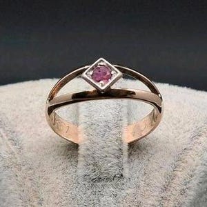 Könnte beinhalten: Ein roségoldener Ring mit geteiltem Schaft-Design. Der Ring hat einen quadratisch geschliffenen, rosa Edelstein, der in einer weißen Metallfassung gefasst ist. Der Ring wird auf einer strukturierten, grauen Oberfläche vor dunklem Hintergrund präsentiert.
