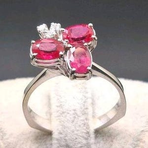 Könnte beinhalten: Ein silberner Ring mit drei ovalen, leuchtend roten Edelsteinen, die sich zu einem Cluster formen, und kleinen, klaren Steinen oben. Der Ring wird auf einer weißen, strukturierten Oberfläche vor dunklem Hintergrund präsentiert.