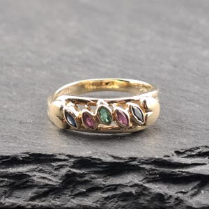 Könnte beinhalten: Ein goldener Ring mit einer Reihe von marquiseförmigen Edelsteinen in Blau, Rosa, Grün und Schwarz. Der Ring hat ein glattes, poliertes Band. Der Ring ist vor einem dunklen Hintergrund platziert.