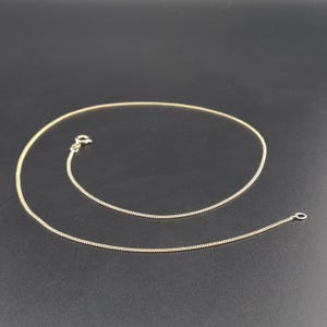 May include: A thin gold chain necklace featuring a delicate curb link design. The necklace is equipped with a small, circular spring ring clasp at one end and a matching jump ring at the other, forming a continuous, smooth, and flexible metallic loop.