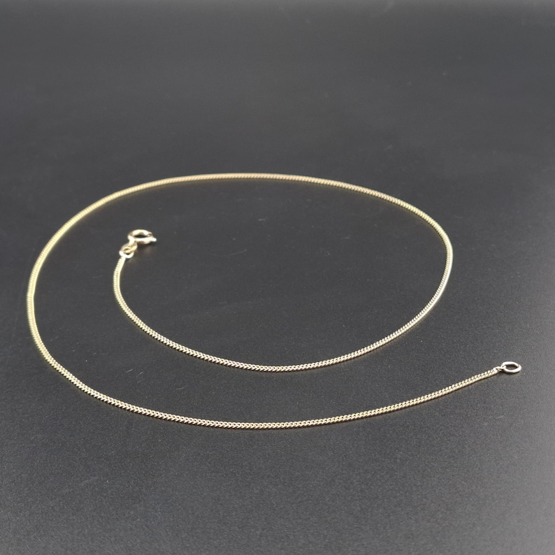May include: A thin gold chain necklace featuring a delicate curb link design. The necklace is equipped with a small, circular spring ring clasp at one end and a matching jump ring at the other, forming a continuous, smooth, and flexible metallic loop.
