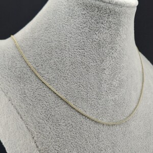 May include: A thin, gold-colored chain necklace featuring a delicate, tightly woven link design. The necklace rests in a smooth, continuous curve, showcasing a polished metallic finish that reflects light along its slender, uniform structure.
