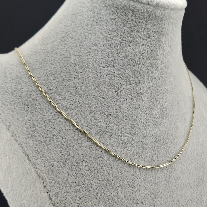 May include: A thin, gold-colored chain necklace featuring a delicate, tightly woven link design. The necklace rests in a smooth, continuous curve, showcasing a polished metallic finish that reflects light along its slender, uniform structure.