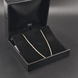 May include: A thin gold chain necklace displayed inside a black jewelry box. The box features a textured exterior and a black velvet interior lining that holds the delicate metal chain in place, showcasing its fine link design and metallic luster.