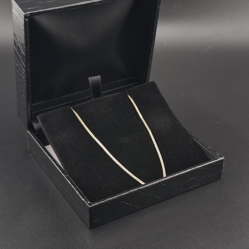 May include: A thin gold chain necklace displayed inside a black jewelry box. The box features a textured exterior and a black velvet interior lining that holds the delicate metal chain in place, showcasing its fine link design and metallic luster.