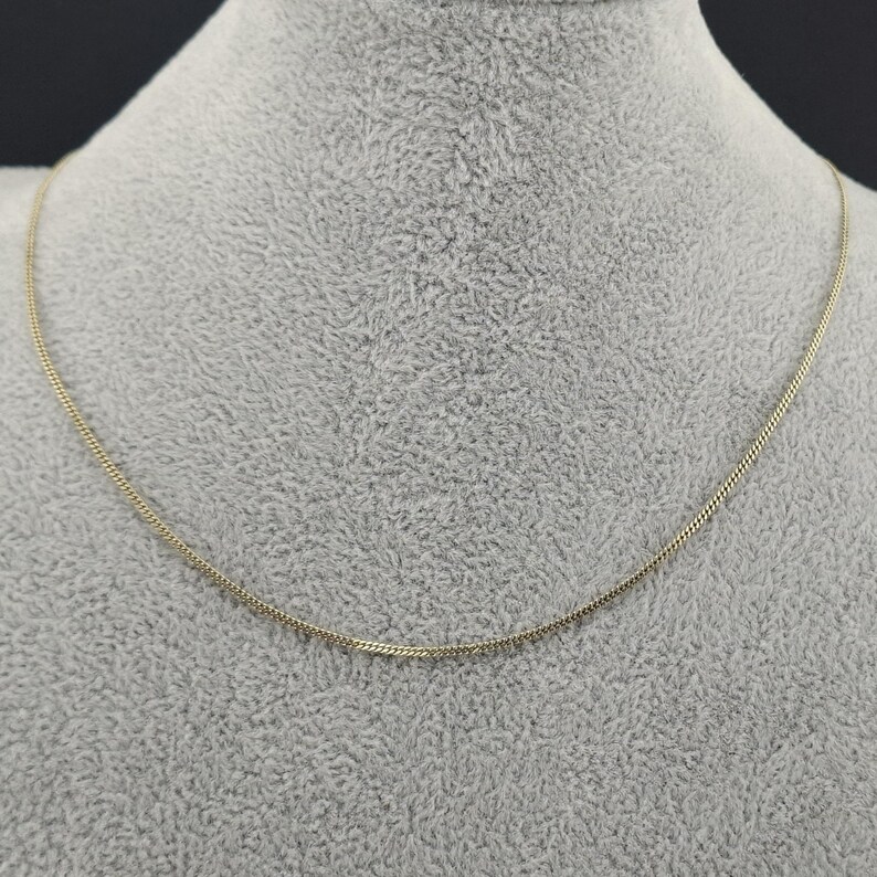 May include: A thin, gold-colored chain necklace featuring a delicate, tightly woven link design. The necklace forms a smooth, continuous curve, showcasing a polished metallic finish that reflects light evenly across its entire length.