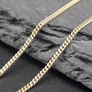 May include: A gold-toned metal chain necklace featuring a classic curb link design. The chain consists of small, uniform, interlocking oval links that create a continuous, flexible strand with a polished finish and a consistent metallic texture.