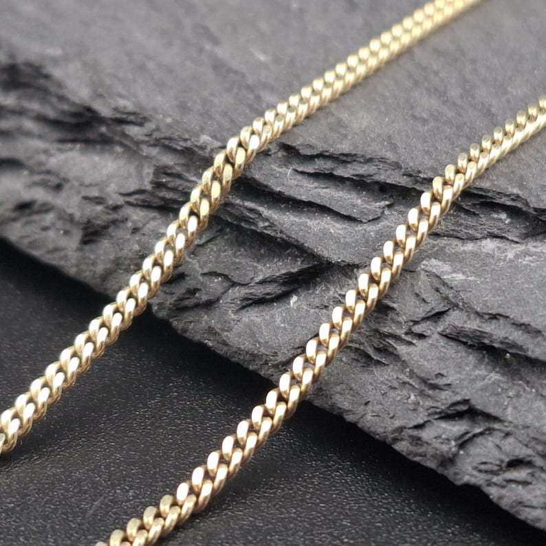 May include: A gold-toned metal chain necklace featuring a classic curb link design. The chain consists of small, uniform, interlocking oval links that create a continuous, flexible strand with a polished finish and a consistent metallic texture.