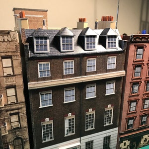 Op de afbeelding: Een model van een drie verdiepingen tellend bakstenen gebouw met witte versieringen en zwarte ramen. Het gebouw heeft een plat dak met drie dakkapellen. Het gebouw is geschilderd in tinten bruin, wit en zwart.