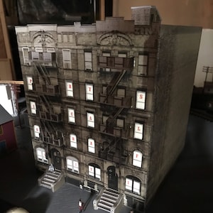 May include: A model of a brick building with fire escapes and a sign that reads "PHYSICAL" on the front. The building is made of cardboard and has a realistic appearance. There are two small figures standing in front of the building.