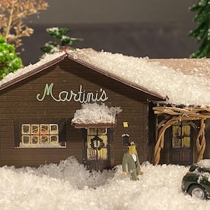 May include: Miniature model of a building with the word "Martini's" in green script above the entrance. The building is covered in snow, with a small figure and a vintage car in front. The scene evokes a winter setting.