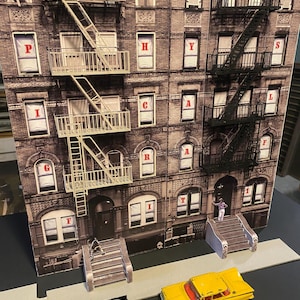 Op de afbeelding: Een diorama van een bakstenen gebouw met brandtrappen en de woorden "Physical Graffiti" in rode letters in de ramen. Twee speelgoedauto's, een zwarte en een gele, staan ​​geparkeerd voor het gebouw.