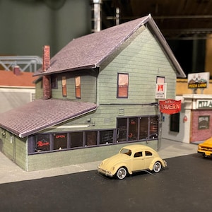 May include: A model train set with a two-story building with a sign that reads "Madison Tavern", a smaller building with a sign that reads "Pizza Land", a yellow pickup truck, and a yellow Volkswagen Beetle.