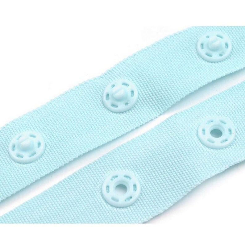 May include: Light blue fabric with plastic snap buttons. The fabric is woven and has a subtle texture. The buttons are round and have a raised centre.