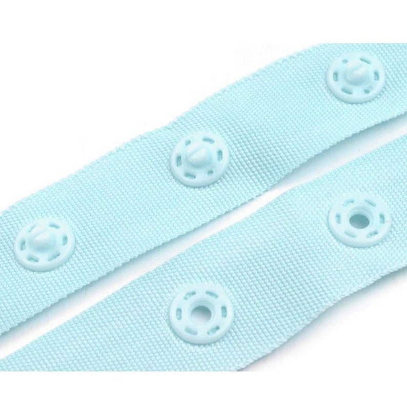 May include: Light blue fabric with plastic snap buttons. The fabric is woven and has a subtle texture. The buttons are round and have a raised center.