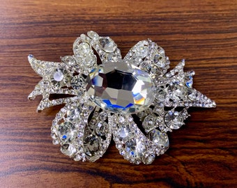 May include: A silver brooch featuring a large, clear crystal at its centre. Smaller crystals surround the main crystal, forming a bow shape.