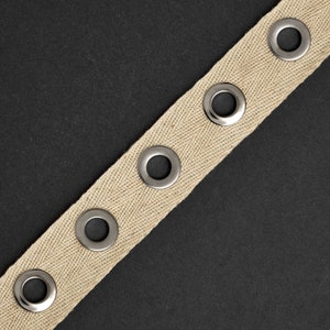 May include: A beige cotton webbing with silver metal eyelets. The webbing is on a black background.