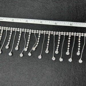 May include: Silver rhinestone fringe trim with a chain of rhinestones and a drop of rhinestones at the end of each fringe. The trim is measured on a ruler with inches.