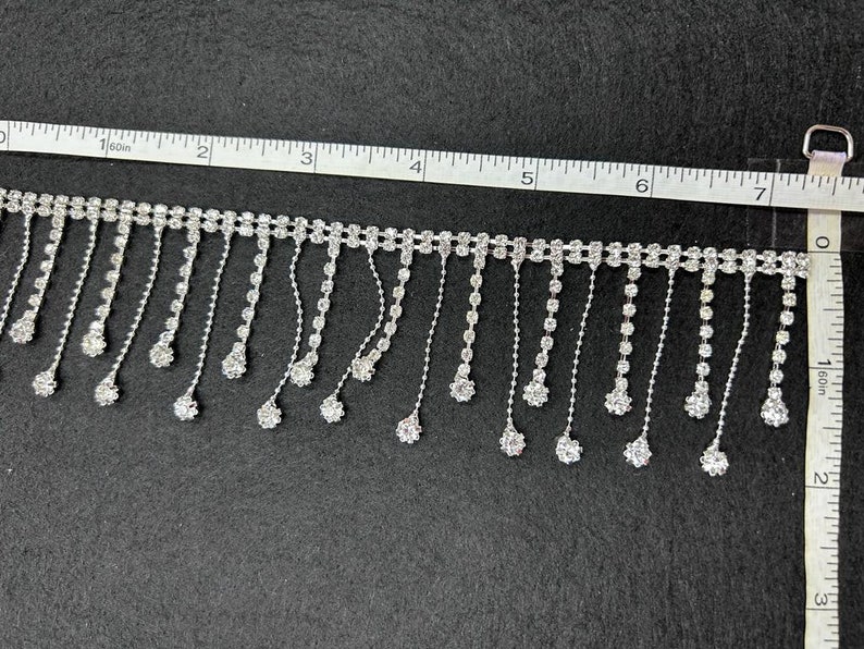 May include: Silver rhinestone fringe trim with a chain of rhinestones and a drop of rhinestones at the end of each fringe. The trim is measured on a ruler with inches.