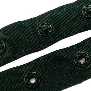 May include: Close-up of dark green fabric with black plastic buttons. The fabric has a woven texture and the buttons have a raised ring around the centre hole.