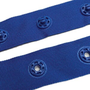 May include: Blue webbing with plastic snap fasteners. The webbing is a woven fabric with a textured surface. The fasteners are round with a small screw in the centre.