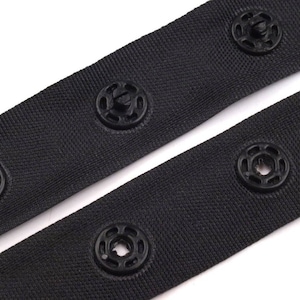 May include: Black fabric tape with black plastic snap buttons. The tape is folded in half, showing the buttons on both sides.