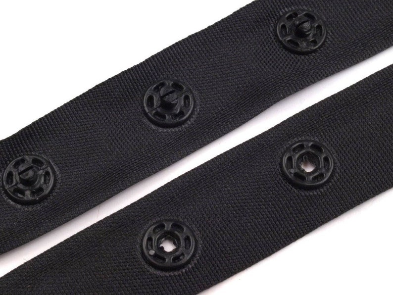 May include: Black fabric tape with black plastic snap buttons. The tape is folded in half, showing the buttons on both sides.