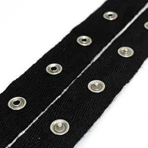 May include: Black fabric ribbon with a white line down the center. The ribbon has silver metal eyelets along the edges.
