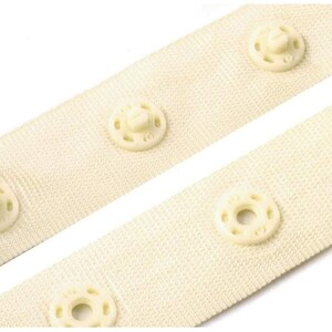 May include: Close-up of two white fabric strips with plastic snap buttons. The strips are sewn together with the buttons facing each other.