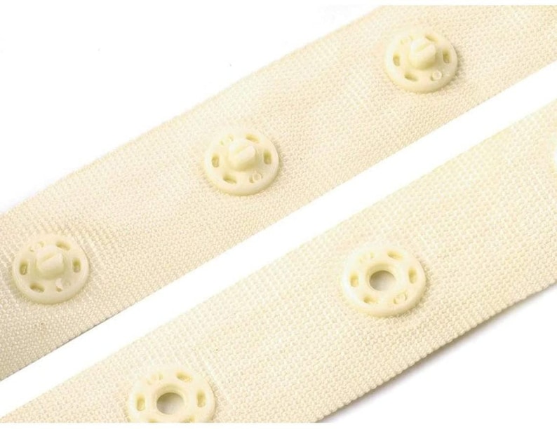 May include: Close-up of two white fabric strips with plastic snap buttons. The strips are sewn together with the buttons facing each other.