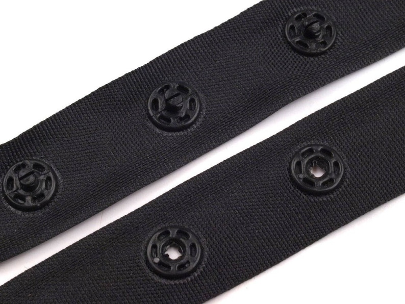 May include: Black fabric tape with black plastic snap buttons. The tape is folded in half, showing the buttons on both sides.