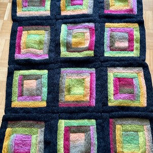 Könnte beinhalten: Handgestrickte Decke mit einem sich wiederholenden Muster aus bunten Quadraten. Jedes Quadrat hat ein zentrales Rechteck, das von konzentrischen Rändern in Rosa-, Grün-, Gelb- und Brauntönen umgeben ist, vor einem dunkelgrauen Hintergrund. Die Decke ist aus weichem Garn.