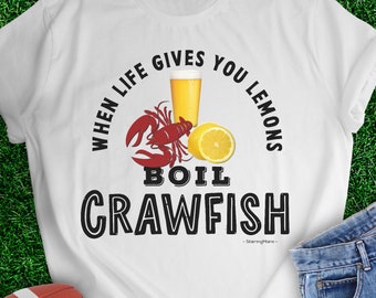 May include: White t-shirt with the text "When life gives you lemons boil crawfish" in black. The shirt also features a red crawfish, a glass of beer, and a lemon.