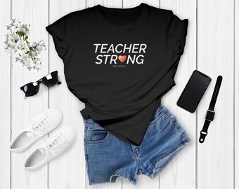 May include: Black t-shirt with white text that reads "TEACHER STRONG" with a red heart graphic. The shirt is laying on a white wooden surface with a pair of sunglasses, white sneakers, denim shorts, a black smartphone, and a black smartwatch.