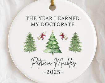 May include: A white ceramic Christmas ornament with a gold string, displayed in a white box. The ornament features text that reads "THE YEAR I EARNED MY DOCTORATE" and the name "Patricia Mackles" with the year "-2025-". It has watercolor illustrations of Christmas trees and bells.